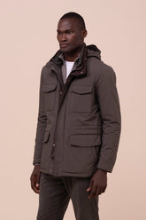 A man wearing a dark olive brown hooded utility jacket with multiple front pockets, standing against a solid beige background.